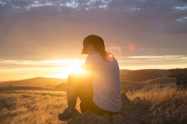 backside of a woman looking down and sitting down.  She's sitting on the ground and the sun is in front of her 